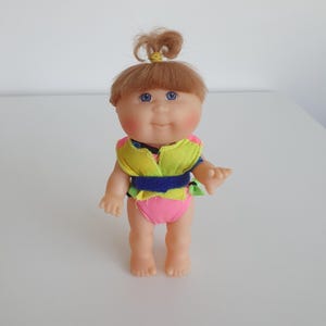 May include: A small doll with light brown hair tied in a top knot, blue eyes, and rosy cheeks. The doll wears a bright neon yellow and pink sleeveless top with a dark blue horizontal strap across the waist and matching pink shorts.