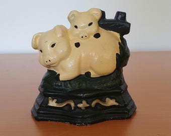 Op de afbeelding: Een deurstop van gietijzer met twee varkensfiguren. Het grotere varken ligt met een kleiner varken op zijn rug. Beide varkens zijn in een lichte gele kleur geverfd. De basis van de deurstop is zwart met een groen en gouden ontwerp.