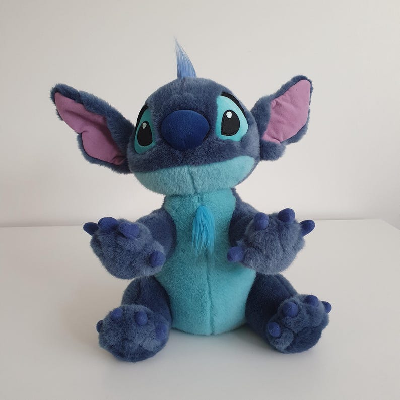 May include: A plush Stitch doll in shades of blue and teal. The doll has large ears with pink inner ears, big black eyes, and a tuft of blue hair. It is sitting with its arms and legs outstretched, showcasing its four-fingered hands and feet.