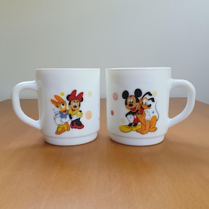 May include: Two white ceramic mugs with cartoon character designs. One mug features Daisy Duck and Minnie Mouse, the other features Mickey Mouse and Pluto. Each mug has a curved handle and colorful illustrations.