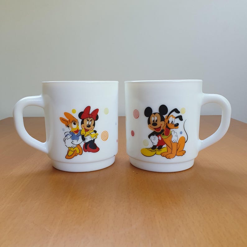 May include: Two white ceramic mugs with cartoon character designs. One mug features Daisy Duck and Minnie Mouse, the other features Mickey Mouse and Pluto. Each mug has a curved handle and colorful illustrations.