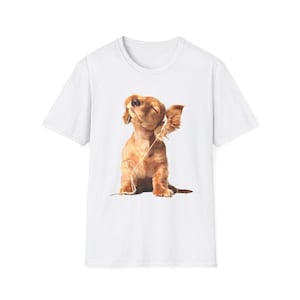 以下が含まれることがあります： 白いTシャツ。茶色のダックスフントの子犬がヘッドホンをしています。子犬は目を閉じており、音楽を聴いているようです。Tシャツはクルーネックです。