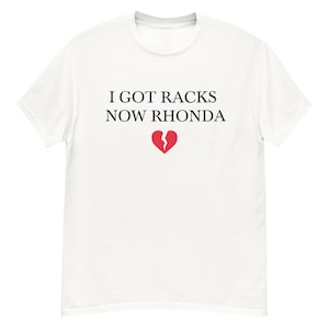 May include: White t-shirt with black text that reads "I got racks now Rhonda" and a red broken heart graphic.