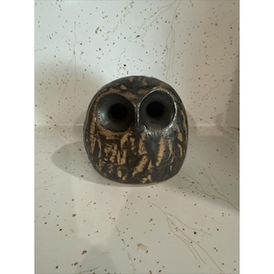 May include: A rounded ceramic owl figurine featuring a dark brown and tan mottled glaze. The sculpture has two large, deep-set circular eye sockets and a smooth, curved body shape with a textured, earthy finish across the entire surface.