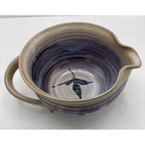 May include: A handmade ceramic mixing bowl with a handle and pouring spout. The bowl features a beige exterior and a blue and purple interior with a painted leaf design. The bowl is ideal for mixing ingredients.