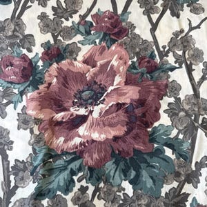 May include: Close-up of a floral fabric featuring large, detailed blooms in shades of burgundy, mauve, and cream. The design includes green leaves and gray branches, creating a vintage, botanical pattern. The fabric has a soft, textured appearance.