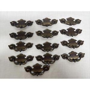 May include: A collection of antique-style drawer pulls. Each pull is made of bronze-colored metal, featuring a decorative, ornate design with a curved handle. The pulls are arranged on a white surface.
