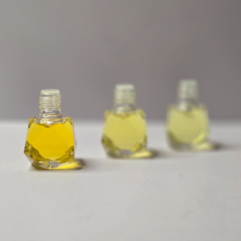 Puede incluir: Tres botellas de vidrio transparente con forma geom&eacute;trica, llenas de un l&iacute;quido amarillo claro. Las botellas est&aacute;n dispuestas en fila sobre una superficie blanca.