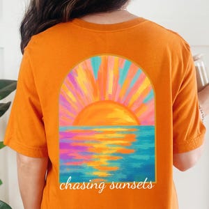 May include: Orange t-shirt featuring a sunset design. The graphic shows a vibrant orange sun setting over a blue and purple ocean, framed by an arch. The words "chasing sunsets" are written below the image.