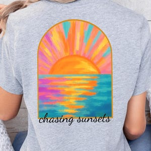 May include: A gray t-shirt with a colorful painting of a sunset over the ocean. The painting is in a gold arch shape. The text 'chasing sunsets' is below the painting.