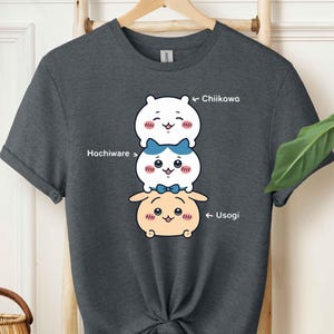 May include: Dark gray t-shirt featuring a stacked cartoon design of three characters with the names Chiikawa, Hachiware, and Usogi. The characters are white, blue, and tan with smiling faces. The shirt is knotted at the waist.