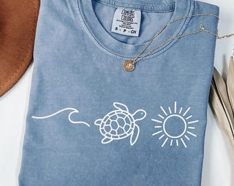 May include: A blue short-sleeved t-shirt with a white graphic of a wave, a turtle, and a sun. The shirt has a crew neck and a small tag on the collar. A gold necklace with a pendant is draped across the shirt.