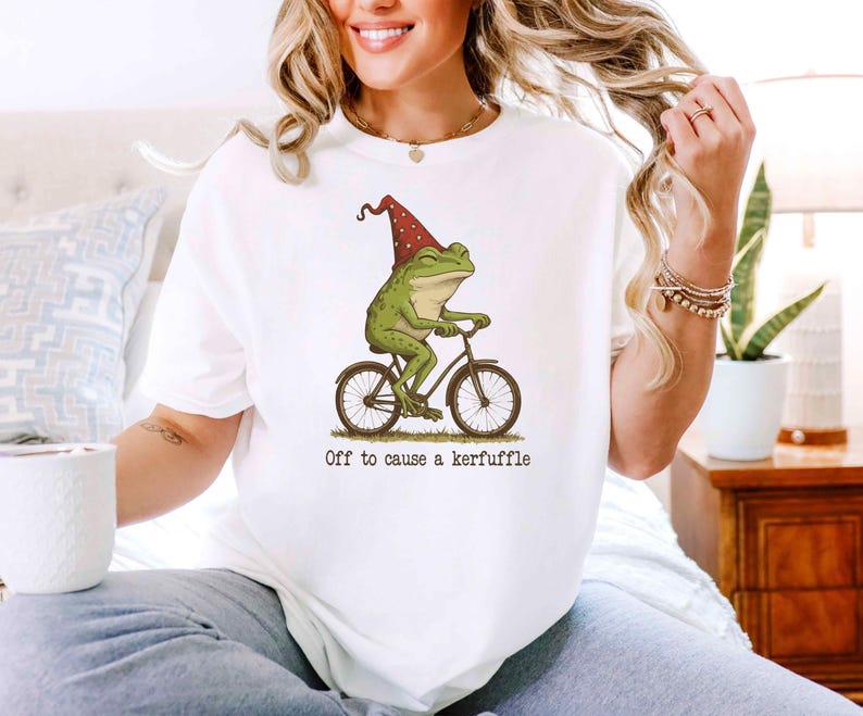 May include: White t-shirt featuring a whimsical illustration of a green frog riding a bicycle and wearing a red and white polka-dot hat. The text "Off to cause a kerfuffle" is printed below the image.
