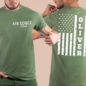 May include: Olive green t-shirt with the text "AIR FORCE Dad" in white on the front. The back of the shirt features a distressed American flag design with the name "OLIVER" printed vertically in white.
