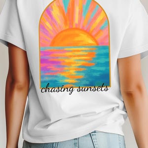 May include: White t-shirt featuring a colorful sunset design with the text "chasing sunsets." The graphic depicts a vibrant orange sun setting over a blue ocean, with pink, orange, and yellow hues. The shirt is a casual wear item.