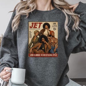 May include: A charcoal gray crewneck sweatshirt featuring a graphic print of a vintage Jet magazine cover from January. The cover displays four individuals with afro hairstyles and the text: JET, THE WEEKLY NEGRO NEWS, JAN, 15C, AFRO GLAMOUR: THE RISE OF GLOBAL STYLES.