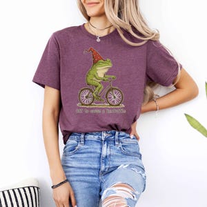 May include: A maroon t-shirt featuring a whimsical illustration of a green frog riding a bicycle and wearing a red hat. The text "Off to cause a little trouble" is below the frog. The shirt is made of soft material.