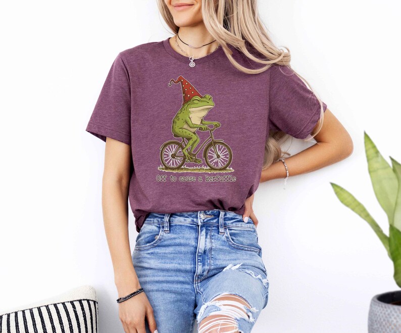 May include: A maroon t-shirt featuring a whimsical illustration of a green frog riding a bicycle and wearing a red hat. The text "Off to cause a little trouble" is below the frog. The shirt is made of soft material.