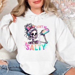 May include: White hoodie with a colorful graphic of a skeleton with a bun in its hair pouring a drink from a bottle. The text "Don't be Salty" is below the skeleton.