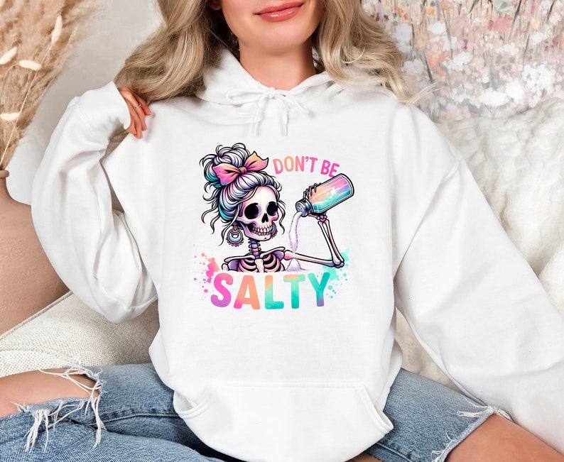 May include: White hoodie with a colorful graphic of a skeleton with a bun in its hair pouring a drink from a bottle. The text "Don't be Salty" is below the skeleton.