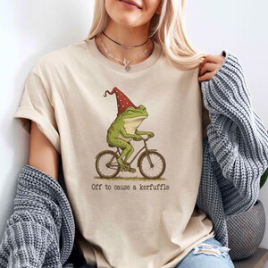 May include: Beige t-shirt featuring a whimsical illustration of a green frog riding a bicycle and wearing a red gnome hat. The text "Off to cause a kerfuffle" is printed below the image. The shirt is made of a soft material.