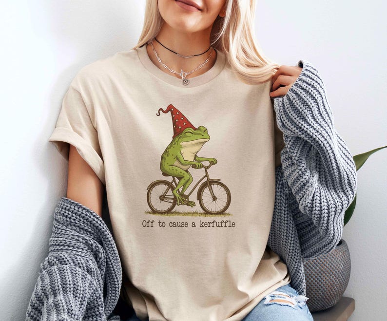 May include: Beige t-shirt featuring a whimsical illustration of a green frog riding a bicycle and wearing a red gnome hat. The text "Off to cause a kerfuffle" is printed below the image. The shirt is made of a soft material.