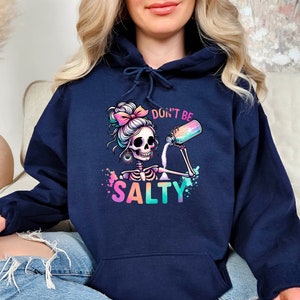 May include: A navy blue hoodie with a colorful graphic of a skeleton with a bun in her hair pouring salt from a shaker. The text "Don't Be Salty" is printed below the graphic.