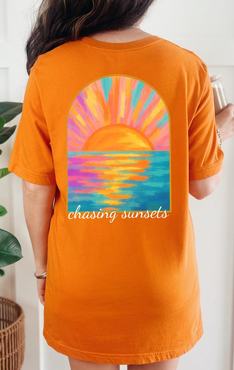 May include: Orange t-shirt featuring a sunset design. The graphic shows a vibrant orange sun setting over a blue and purple ocean, framed by an arch. The words "chasing sunsets" are written below the image.
