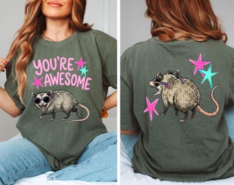 May include: Olive green t-shirt with a graphic of an opossum wearing sunglasses. The front of the shirt says "YOU'RE AWESOME" in pink and teal. The back of the shirt has the same opossum graphic and stars.