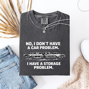 May include: Dark gray t-shirt with the text "NO, I DON'T HAVE A CAR PROBLEM. I HAVE A STORAGE PROBLEM." and a graphic of four classic cars. The shirt is laid flat with blue jeans, white shoes, and a necklace.