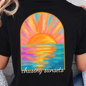 May include: Black t-shirt featuring a colorful sunset design. The graphic shows an orange sun setting over a blue ocean, with the words "chasing sunsets" in a script font. The design is framed by an arch.