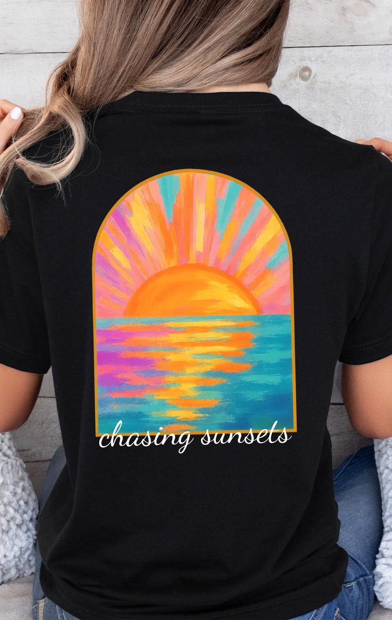 May include: Black t-shirt featuring a colorful sunset design. The graphic shows an orange sun setting over a blue ocean, with the words "chasing sunsets" in a script font. The design is framed by an arch.