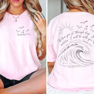 May include: A light pink t-shirt with a black graphic of birds and the text "Isaiah 43:2" on the front. The back features a sun, birds, a wave, and the quote "When you go through deep waters, I will be with you. Isaiah 43:2."