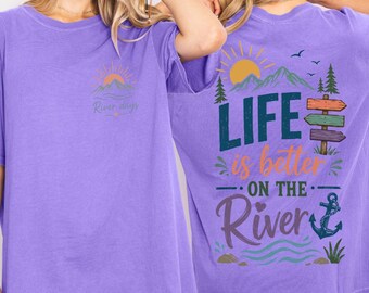 May include: A purple t-shirt featuring a small graphic on the front with a sun, mountains, and the text River days. The back displays a large graphic with mountains, a sun, trees, a signpost, an anchor, and the text LIFE is better ON THE River.