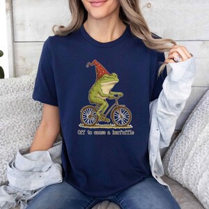 May include: Navy blue t-shirt featuring a whimsical illustration of a green frog riding a bicycle and wearing a red hat. The text "Off to cause a kerfuffle" is below the image. The shirt is made of a soft material.