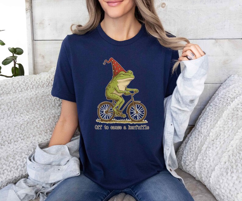 May include: Navy blue t-shirt featuring a whimsical illustration of a green frog riding a bicycle and wearing a red hat. The text "Off to cause a kerfuffle" is below the image. The shirt is made of a soft material.