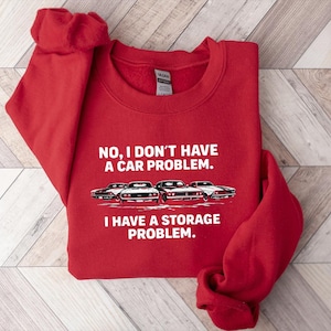 May include: A red sweatshirt with the text "NO, I DON'T HAVE A CAR PROBLEM. I HAVE A STORAGE PROBLEM." and a graphic of four classic cars. The sweatshirt is folded on a light wood surface.