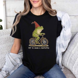 May include: Black t-shirt featuring a whimsical illustration of a green frog wearing a red hat and riding a bicycle. The text "Off to cause a kerfuffle" is printed below the image. The shirt is made of a soft material.