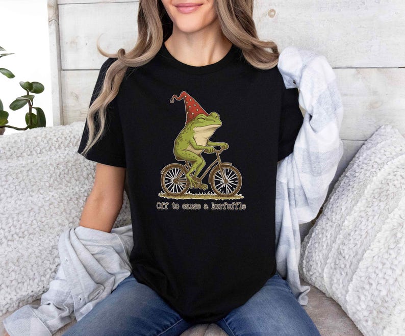 May include: Black t-shirt featuring a whimsical illustration of a green frog wearing a red hat and riding a bicycle. The text "Off to cause a kerfuffle" is printed below the image. The shirt is made of a soft material.