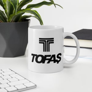 May include: White ceramic mug with a black logo of three stacked T's and the word "TOFAS" printed below.