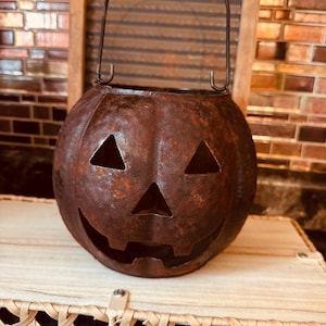 May include: A round, dark brown metal pumpkin bucket with a distressed, rusted finish. It features two triangular eyes, a triangular nose, and a wide, jagged-toothed grin. A thin, dark metal wire handle is attached to the top for carrying.