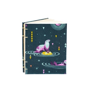 Puede incluir: Un peque&ntilde;o cuaderno abierto con una cubierta de color verde azulado oscuro que presenta un dise&ntilde;o caprichoso de nutria. La ilustraci&oacute;n de la cubierta incluye una nutria rosa y blanca sobre una roca amarilla, rodeada de agua, estrellas y formas geom&eacute;tricas. La encuadernaci&oacute;n es dorada.