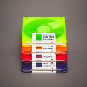 Pu&ograve; includere: Una pila di floppy disk da 3,5 pollici colorati in verde, arancione, rosso e blu. Ogni disco ha un'etichetta bianca con il testo "2HD IBM" e "imation". I dischi sono impilati su una superficie grigia.