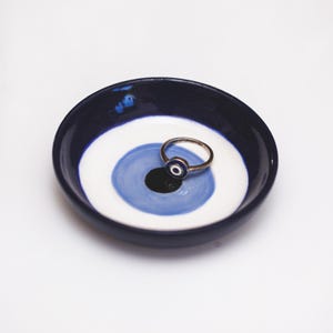 May include: A small blue and white ceramic dish with a blue evil eye design. A gold ring with a blue evil eye is sitting in the dish.