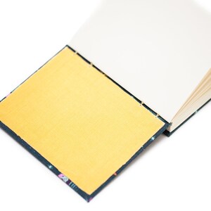 Puede incluir: Un cuaderno abierto con una cubierta dorada y p&aacute;ginas blancas. La cubierta tiene un borde azul oscuro con un patr&oacute;n colorido. El cuaderno est&aacute; sobre un fondo blanco.