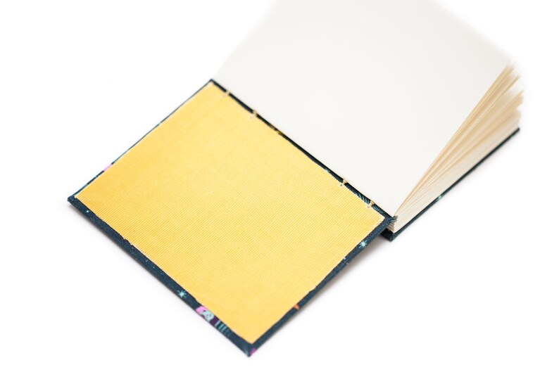 Puede incluir: Un cuaderno abierto con una cubierta dorada y p&aacute;ginas blancas. La cubierta tiene un borde azul oscuro con un patr&oacute;n colorido. El cuaderno est&aacute; sobre un fondo blanco.