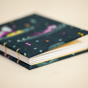 Puede incluir: Un peque&ntilde;o cuaderno abierto con una cubierta de color verde oscuro con un dise&ntilde;o celestial colorido. Las p&aacute;ginas son blancas y el cuaderno est&aacute; encuadernado con una fina banda el&aacute;stica amarilla. El fondo es beige claro.