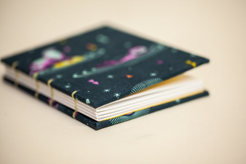 Puede incluir: Un peque&ntilde;o cuaderno abierto con una cubierta de color verde oscuro con un dise&ntilde;o celestial colorido. Las p&aacute;ginas son blancas y el cuaderno est&aacute; encuadernado con una fina banda el&aacute;stica amarilla. El fondo es beige claro.