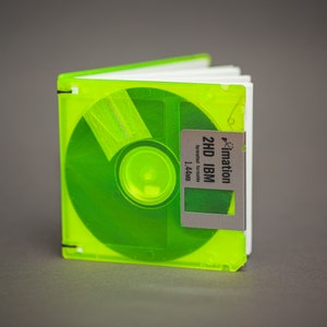 Pu&ograve; includere: Un floppy disk verde neon trasformato in un taccuino. L'etichetta del disco recita "2HD IBM formatted formatted 1.44MB" con il logo Imation. Il taccuino ha pagine bianche e la cerniera del disco originale forma il dorso.