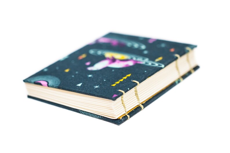 Puede incluir: Un peque&ntilde;o cuaderno de tapa dura con una cubierta azul oscuro que presenta un dise&ntilde;o tem&aacute;tico espacial. La cubierta muestra ilustraciones de planetas, estrellas y naves espaciales en tonos de rosa, morado y amarillo. Las p&aacute;ginas son de color crema y la encuadernaci&oacute;n est&aacute; asegurada con hilo dorado.